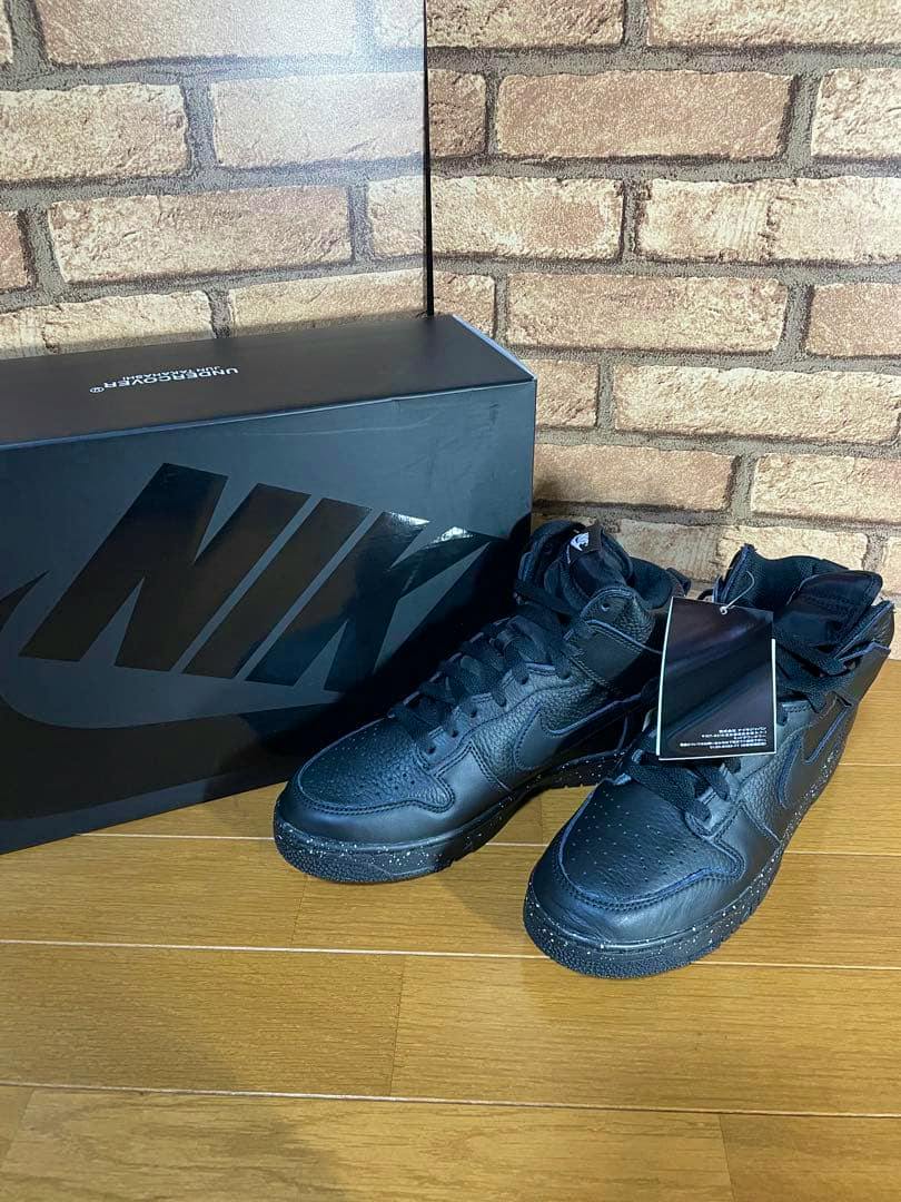 Black Nike Dunk Hi 1985 Undercover sneakers with tags displayed next to a black Nike shoebox on a wooden floor.