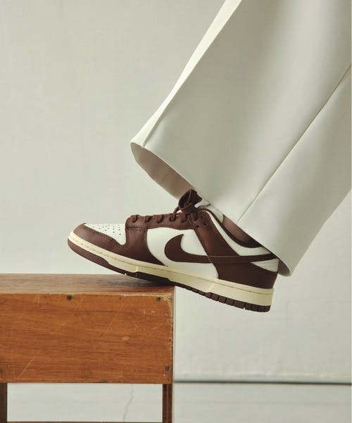 A brown and white NIKE DUNK LOW OG sneaker shown on a foot resting on a wooden bench.