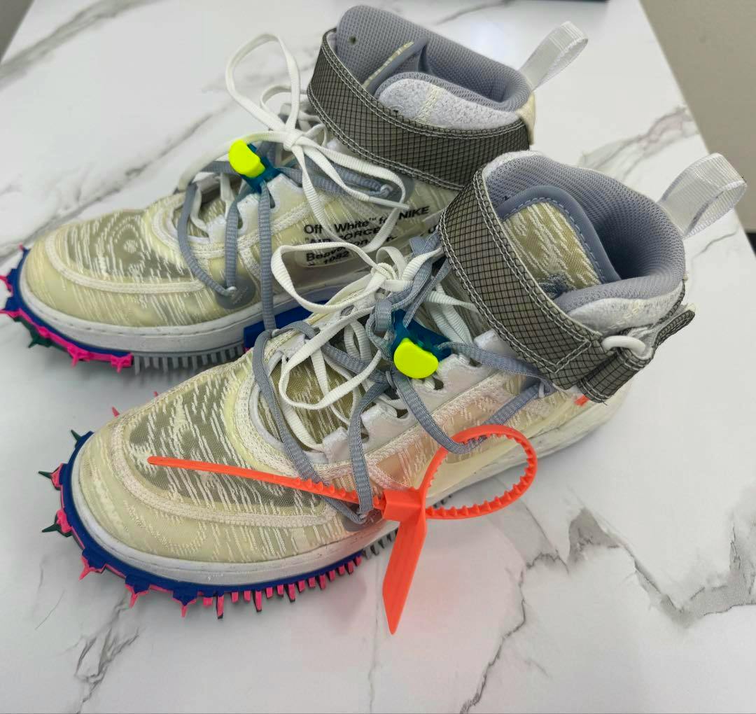 NIKE Off-White AIR FORCE 1 MID sneakers in cream and gray with spiky multicolor soles and an orange zip tie, shown angled on a white surface.