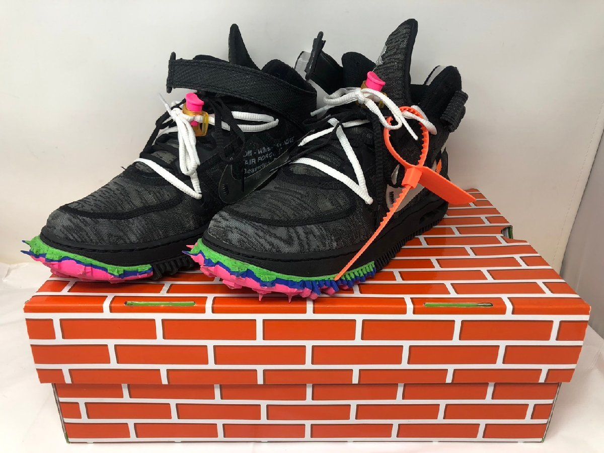 Pair of black Nike × Off-White Air Force 1 Mid sneakers with multicolored soles and an orange zip tie displayed on an orange brick-patterned shoebox.