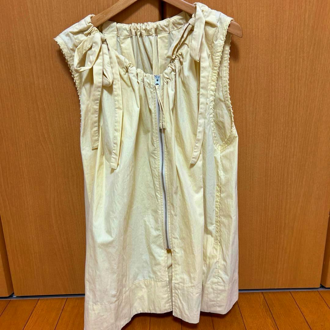 Front view of a light-colored polyester sleeveless zip-front vest hanging on a wooden hanger against a wooden background.