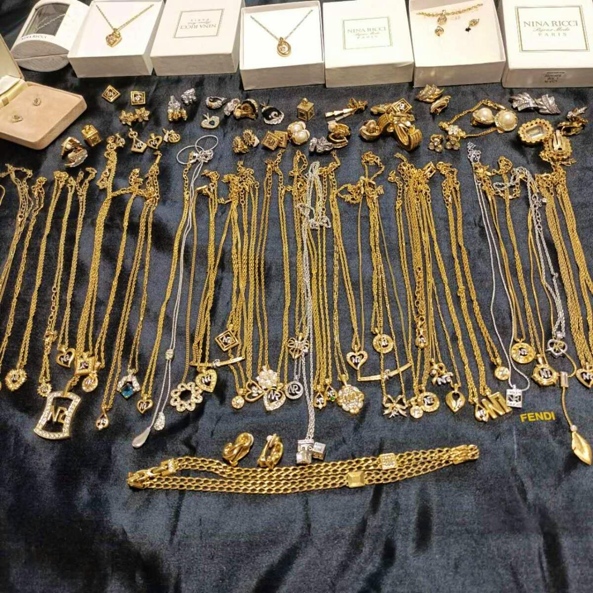 Multiple gold-tone necklaces and pendant charms spread on a dark cloth with several Nina Ricci boxes visible in the background.