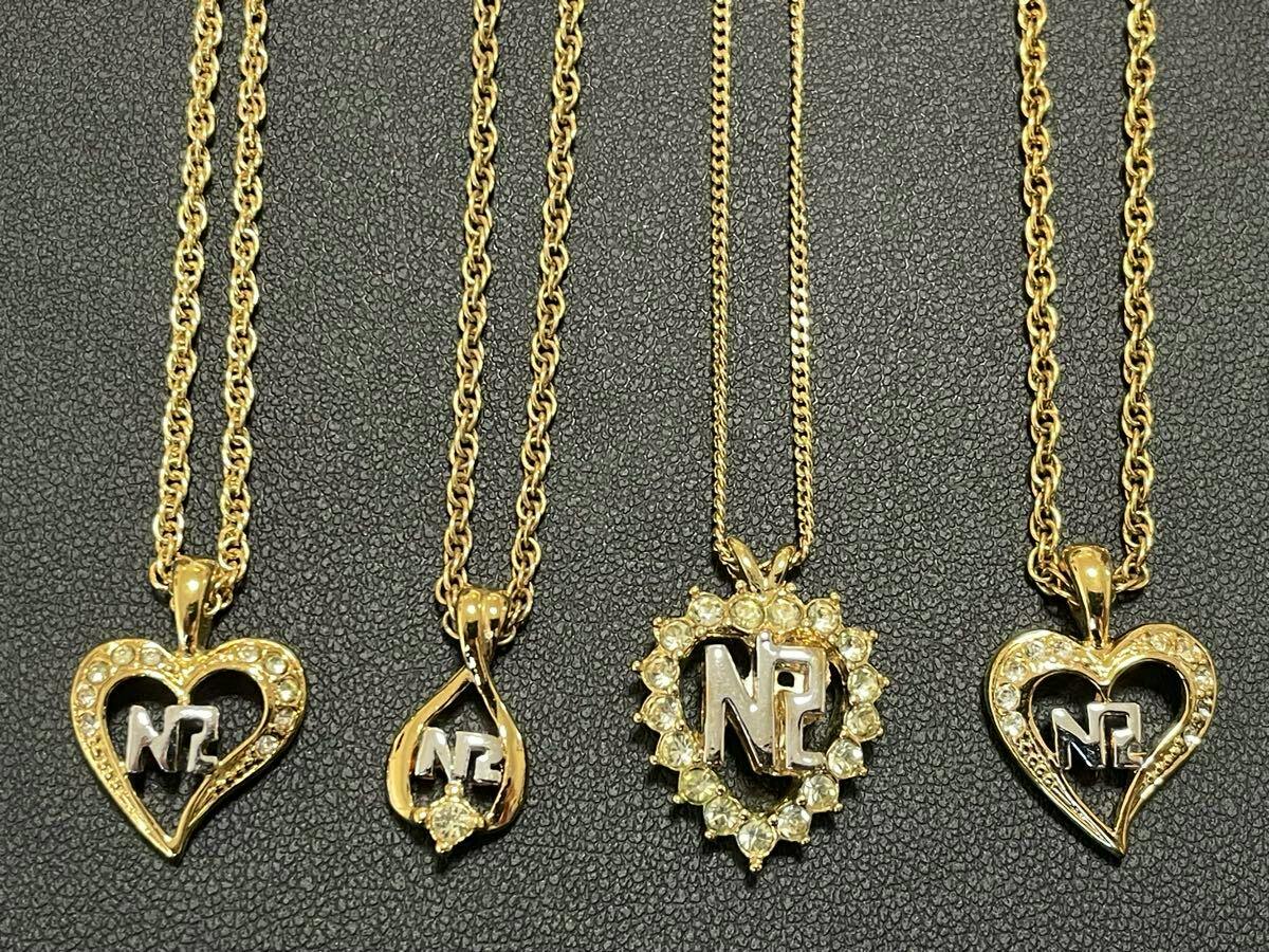 Close-up of four gold-color NINA RICCI necklace pendants with NR logos and rhinestone decorations on a dark textured background.