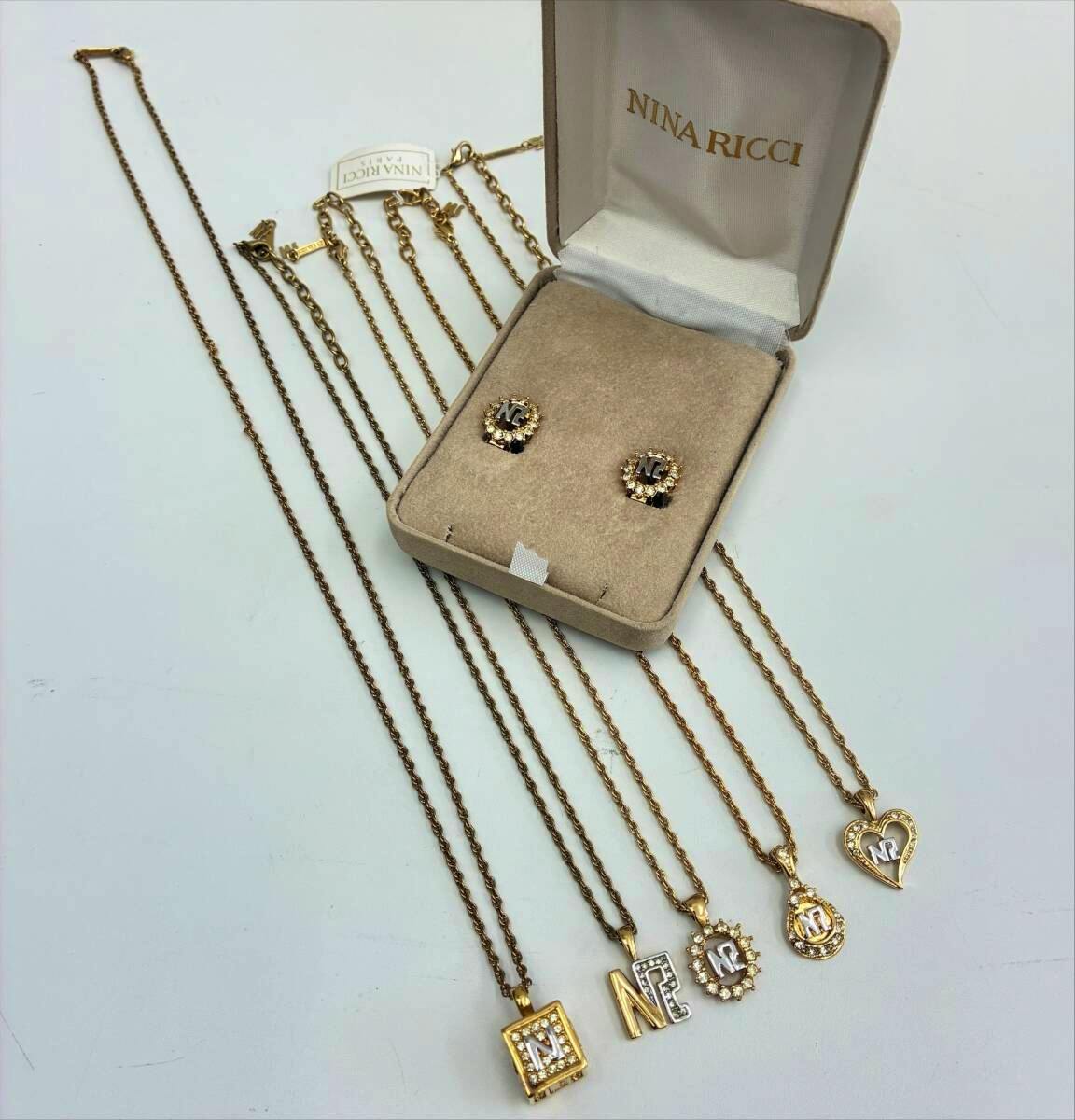 Five gold-colored Nina Ricci necklaces with pendants and a pair of Nina Ricci earrings displayed in an open Nina Ricci jewelry box on a white surface.