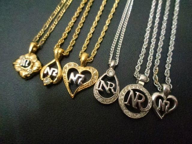 Six NINA RICCI necklaces in gold and silver with NR pendant charms displayed on a dark background.