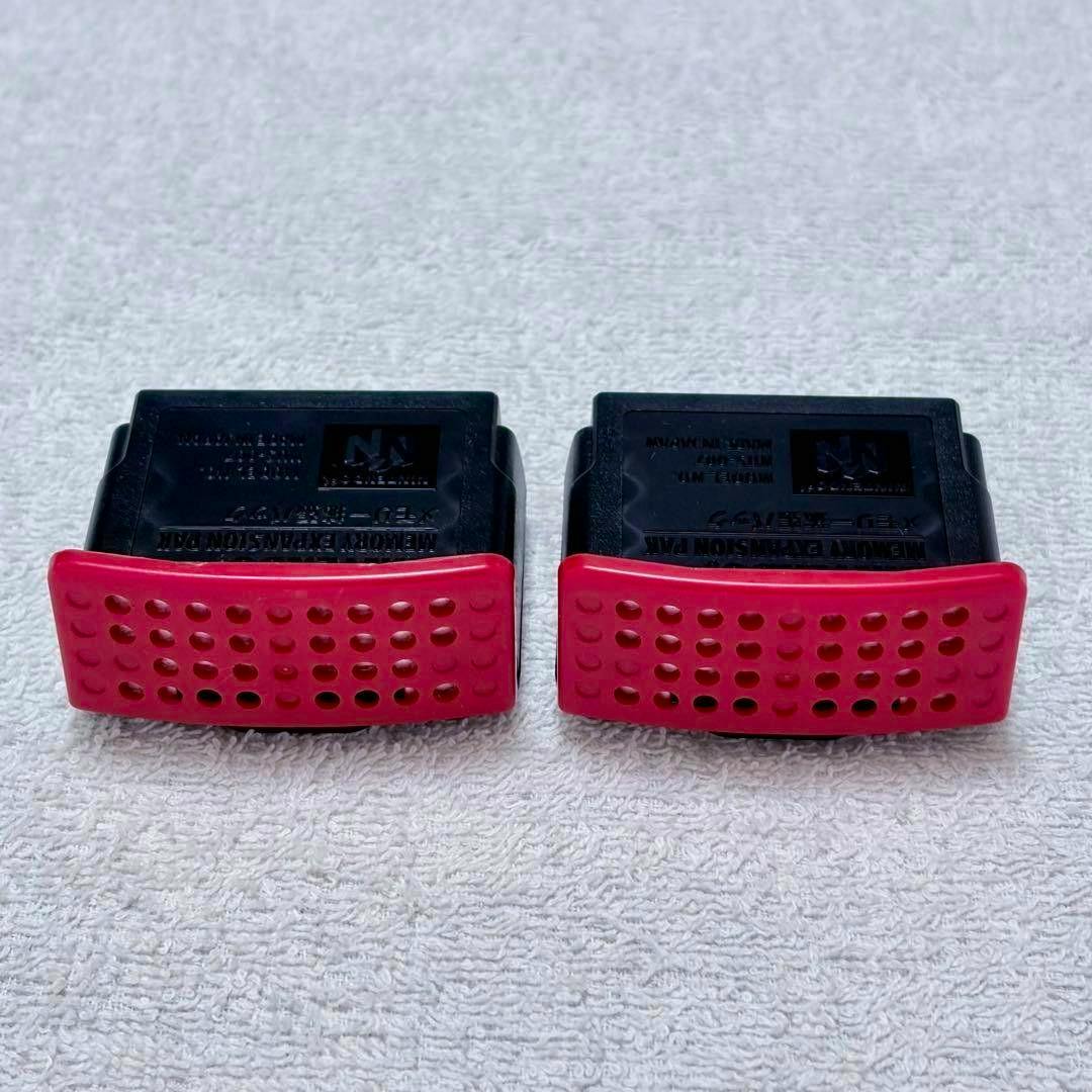 Two Nintendo 64 memory expansion/rumble packs with red perforated fronts placed on a white textured surface.
