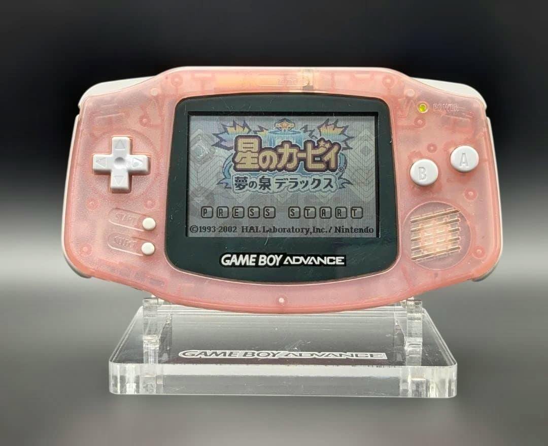 Front view of a milky pink Nintendo Game Boy Advance (AGB-001) on a clear stand showing its screen and controls.