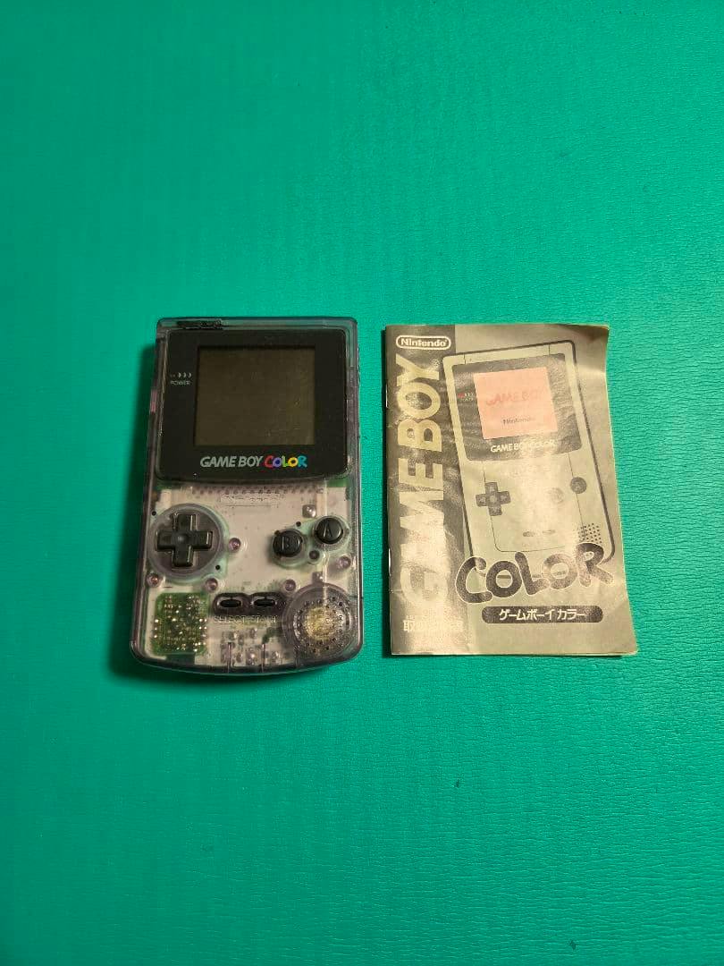 Clear (transparent) Game Boy Color handheld console shown next to its instruction manual on a green surface.