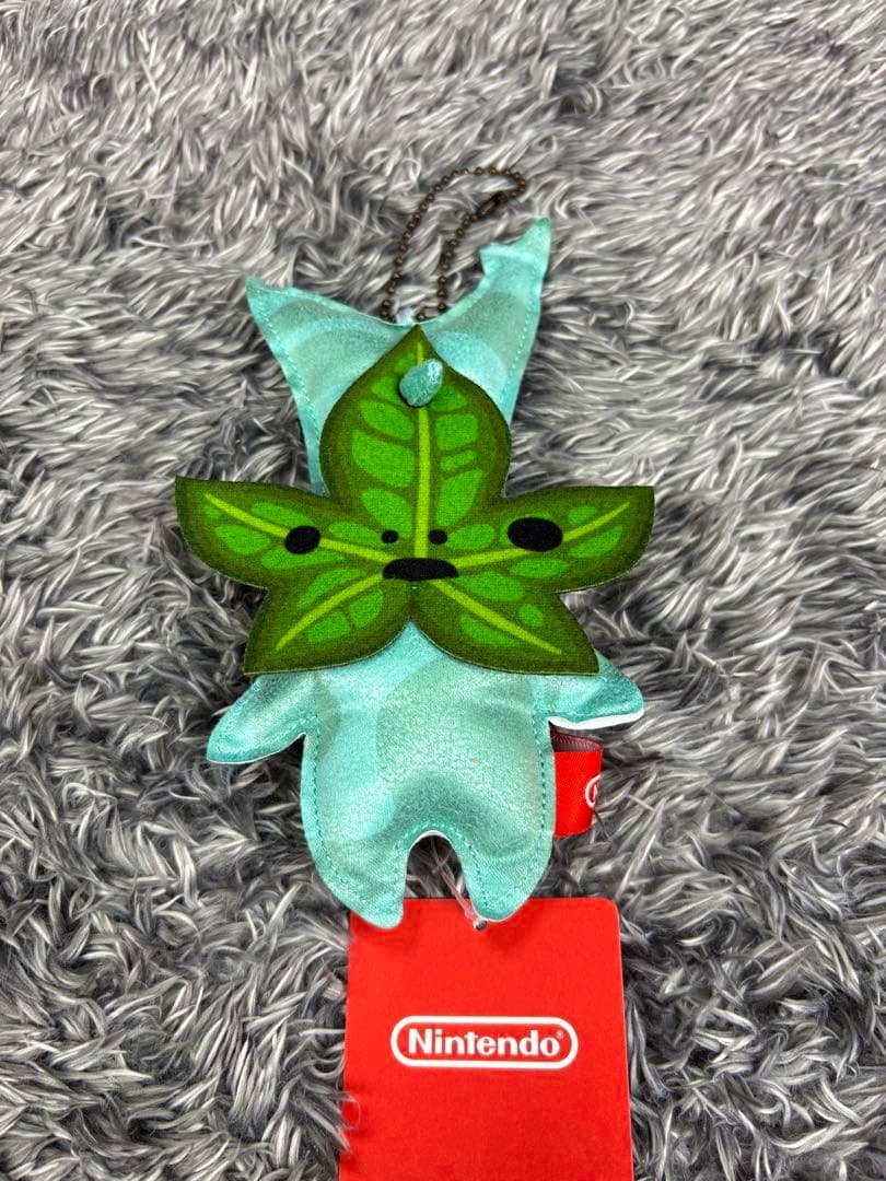 A Korok plush keychain from Zelda with a green leaf face and a red Nintendo tag lying on gray faux fur.