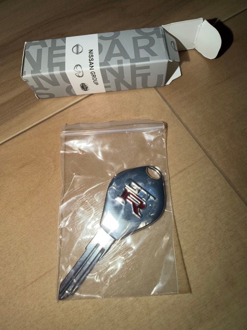 A Nissan BCNR33 Skyline GT‑R blank master key with GT‑R logo in a plastic bag on a wood floor, with a Nissan parts box visible.