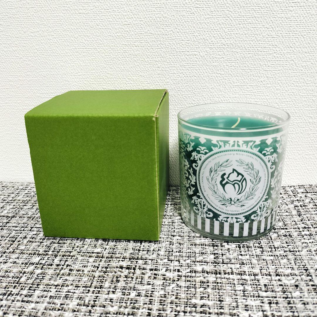 A green aroma candle in a glass container with a white decorative emblem pattern shown next to a plain green box on a textured surface.