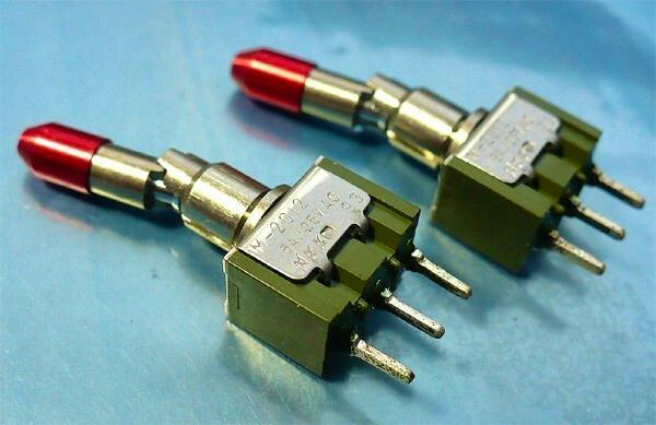 Two NKK M2012 lever-lock toggle switches with red caps placed on a blue surface.