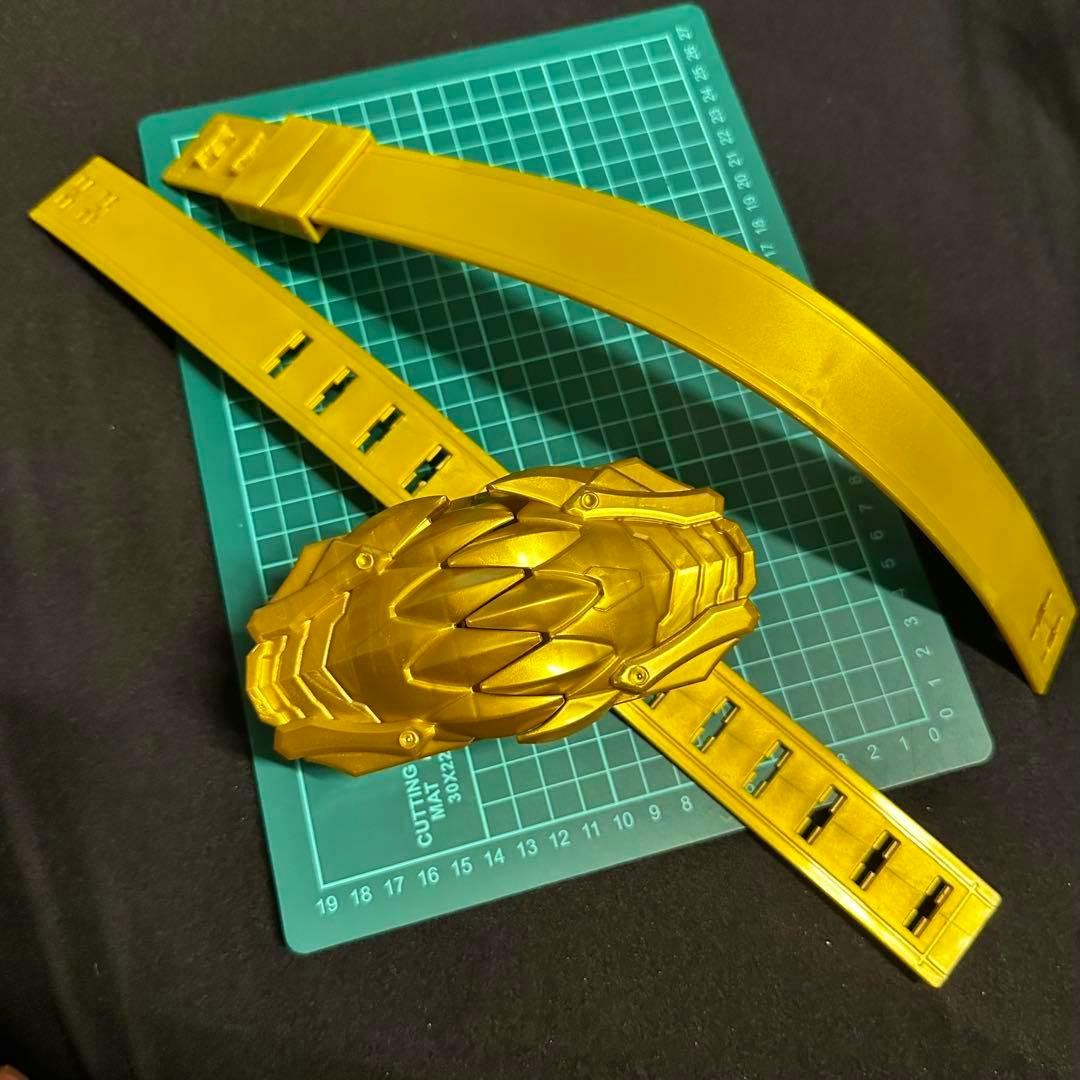 Gold-colored plastic DX Tsumega buckle and two belt strap pieces laid on a green cutting mat.