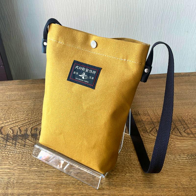 Orange 10号帆布 tablet pochette (canvas pouch) with a black shoulder strap and front label shown on a clear stand on a wooden surface.