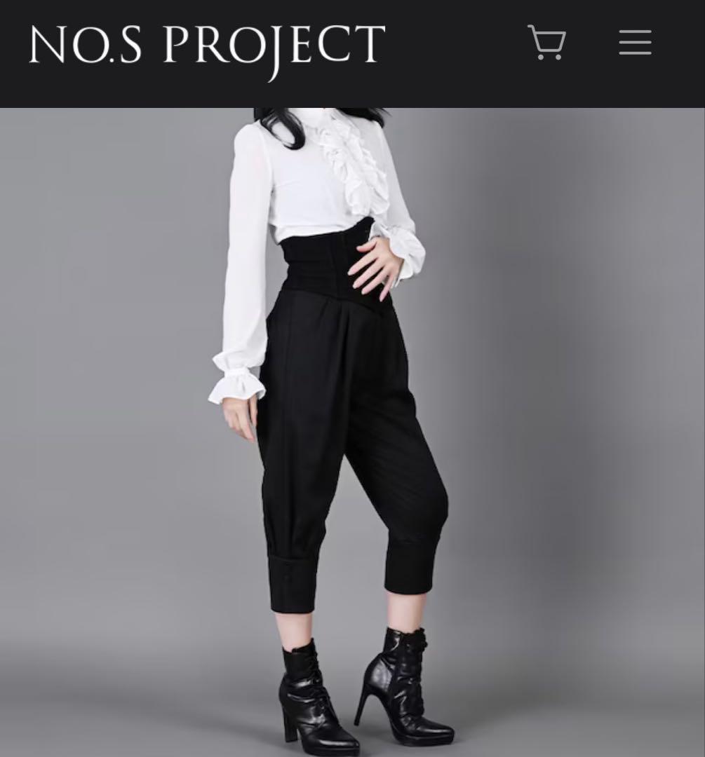 A model wearing black riding-style women’s pants with a high waistband and cropped cuffed legs, styled with a white ruffled blouse and black ankle boots.
