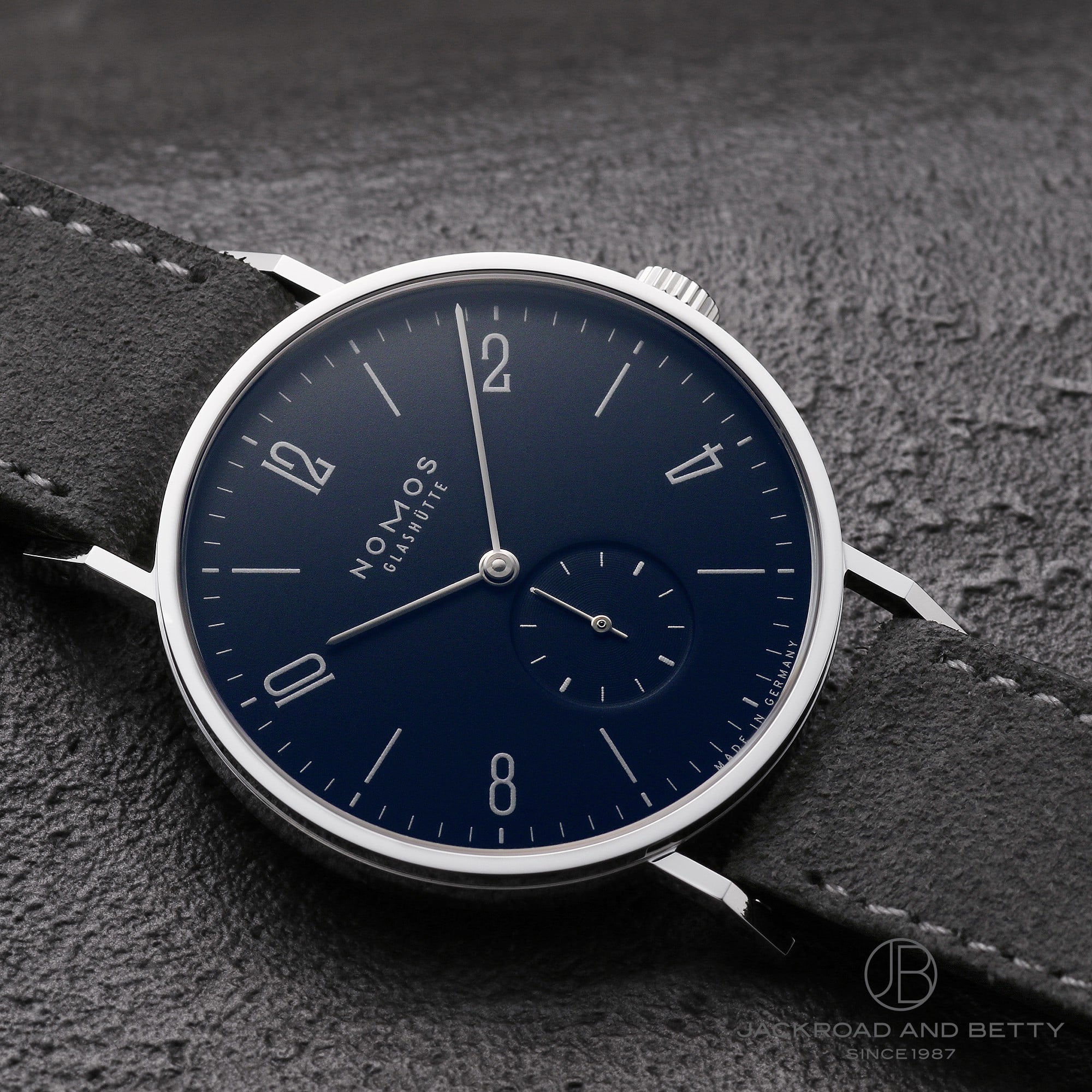 Close-up of the NOMOS Tangent Midnight Blue wristwatch showing its midnight-blue dial, small seconds subdial, metal case, and gray leather strap on a textured background.