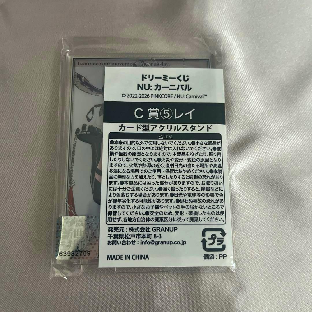 Packaged NU:カーニバル C賞 card-type acrylic stand (Rei) in clear plastic with a Japanese product label visible.