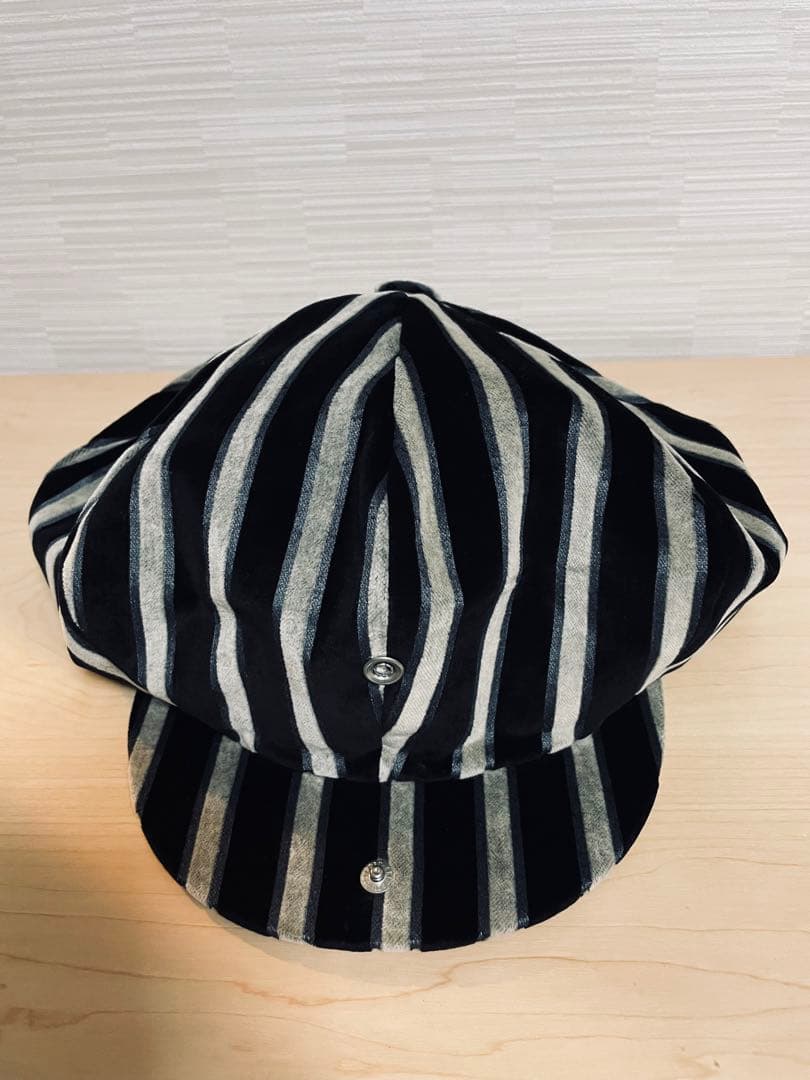 Front view of a striped cotton casquette (hat) with black and gray vertical stripes and a short visor on a light surface.
