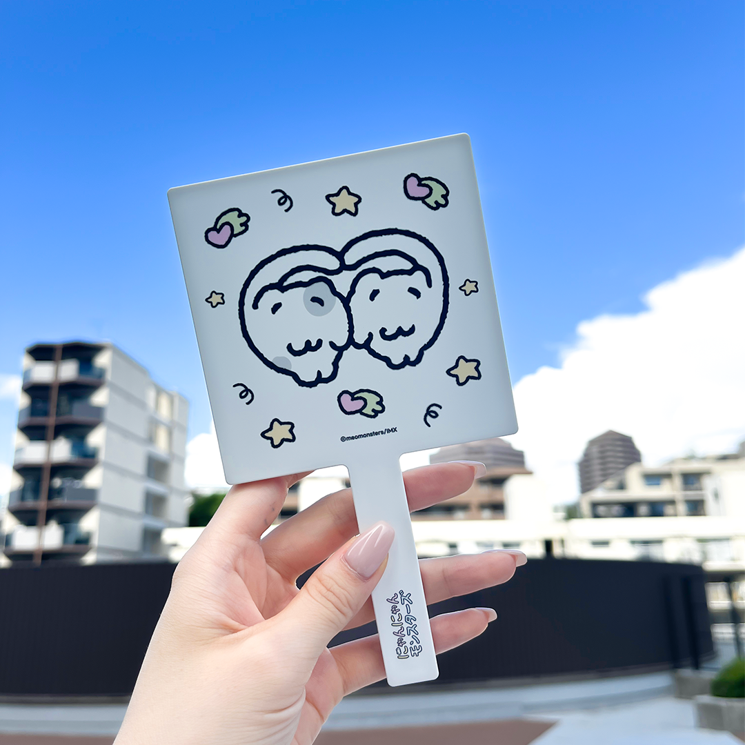 A white handheld compact mirror with a cartoon double-character and star motifs, held by a hand against a blue sky.