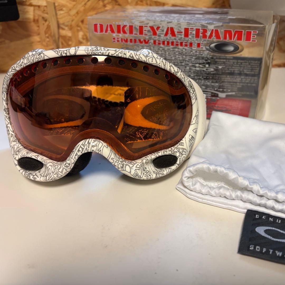 White Oakley A-Frame snow goggles with an orange-tinted lens shown with box and cloth pouch.