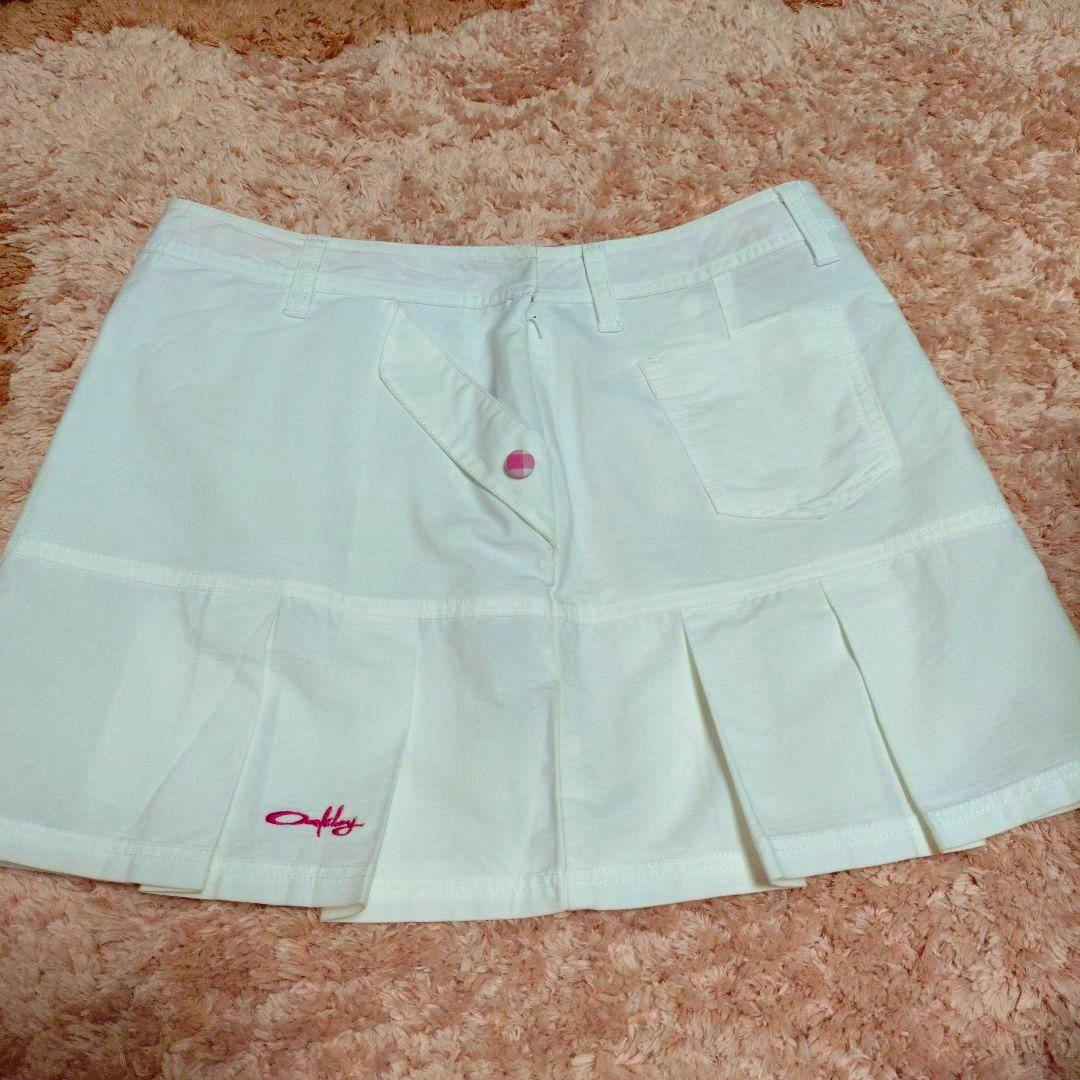 White Oakley golf skirt laid flat on a carpet showing the waistband, pleated hem, back pocket, and pink Oakley logo.