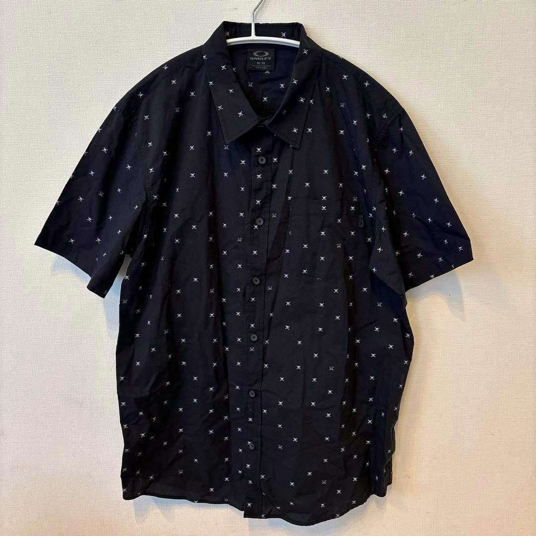 Black Oakley short-sleeve men's button-up shirt with a repeating small skull pattern hanging on a hanger.