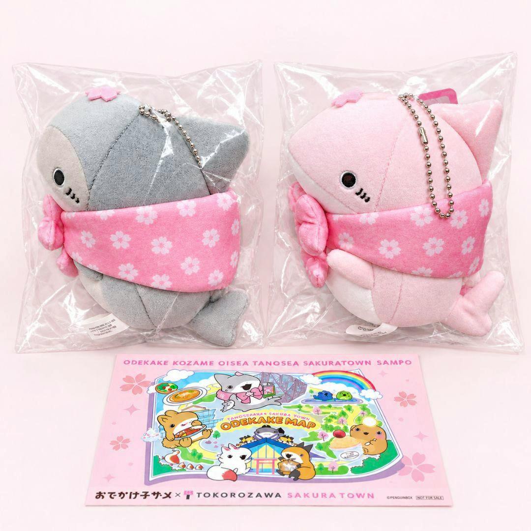Two baby shark plush toys (one gray, one pink) in clear plastic bags wearing pink sakura-patterned scarves, shown above a pink illustrated Odekake map.