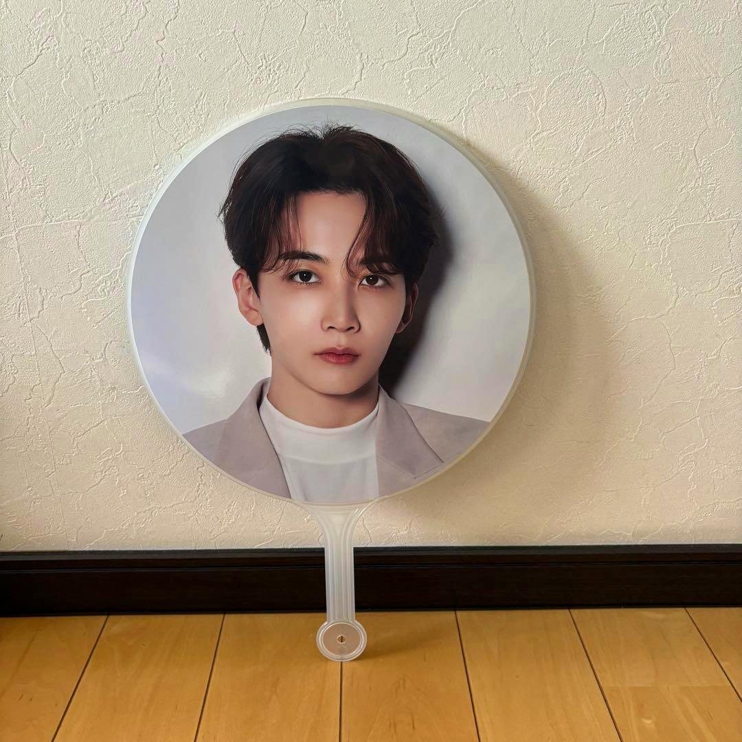 Round paper fan with a printed portrait of a young man attached to a clear plastic handle, photographed leaning against a textured wall.