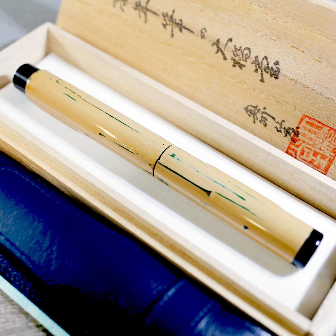 Lacquered ebonite fountain pen with a cream body and green markings displayed in a wooden presentation box with Japanese writing, with a blue case visible.