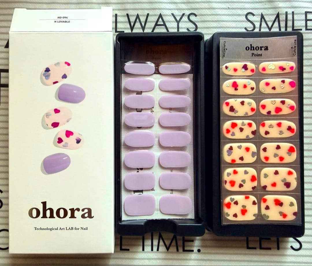 Top-down photo of the ohora N Lovable nail sticker set showing the product box and two trays of stickers: solid lavender nails and cream nails with small red and silver heart designs.