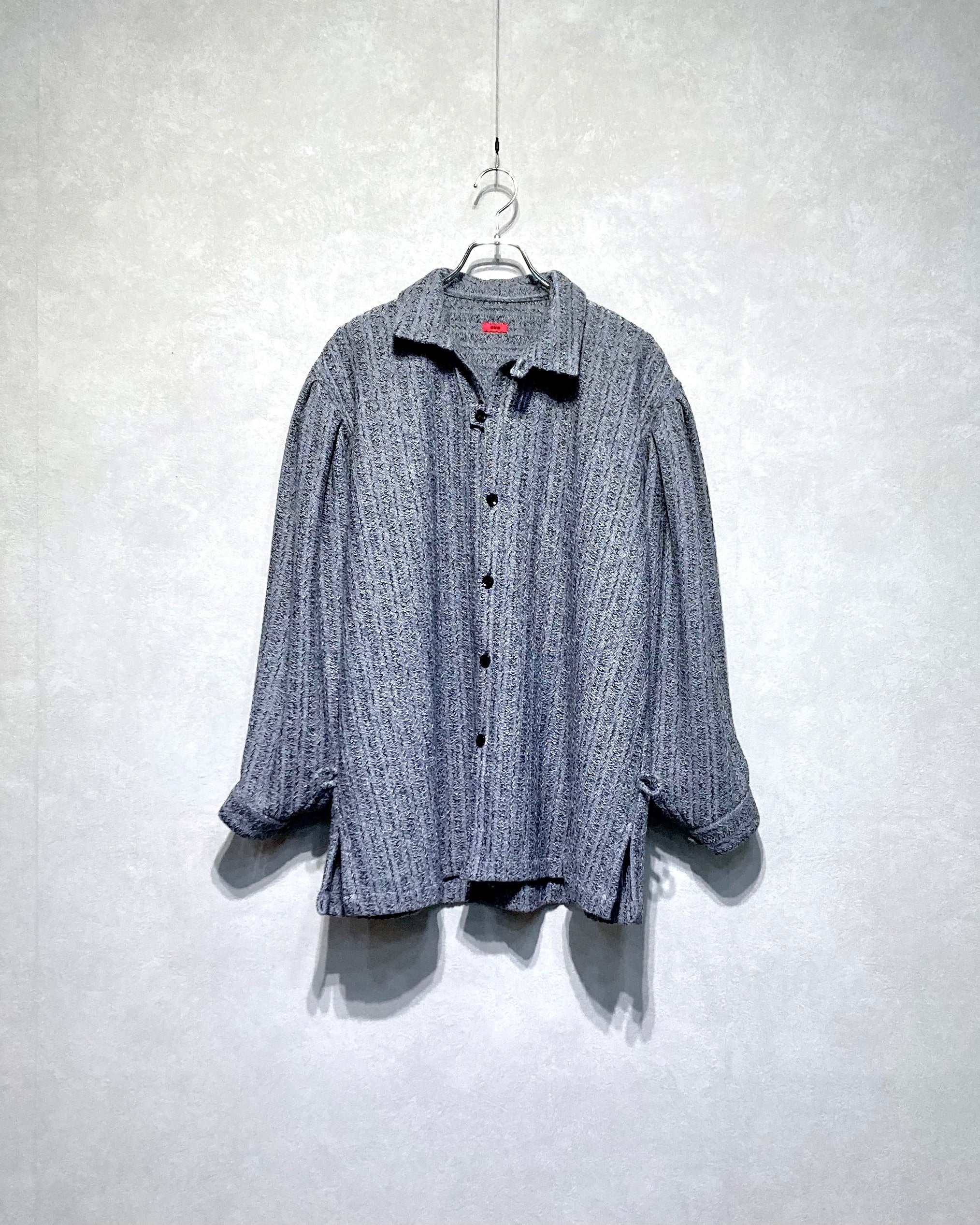 Front view of the strangely woven wool sweater (dress-shirt style) with buttons hanging on a hanger against a light wall.