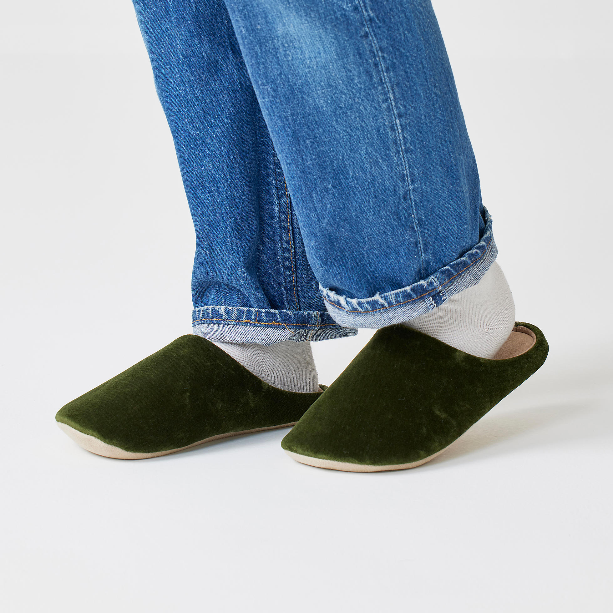 A person wearing the dark green おかげ庵ソファー柄ルームシューズ slippers with white socks and rolled blue jeans.