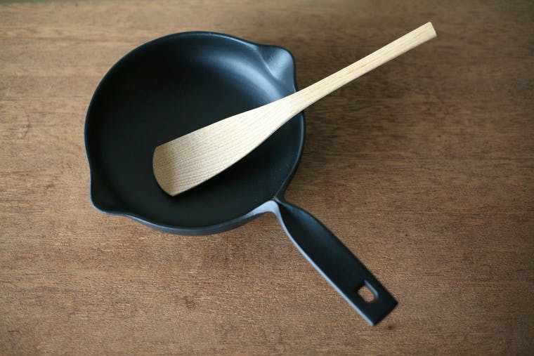 A wooden cooking spoon resting inside a black frying pan on a wooden table.