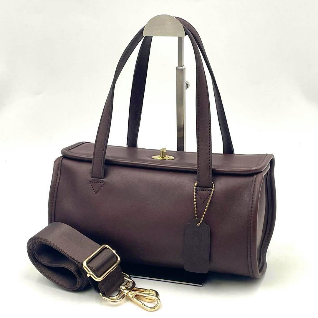 Brown cow-leather 2-way handbag with top handles, a turn-lock closure, and a coiled detachable shoulder strap on a display stand.