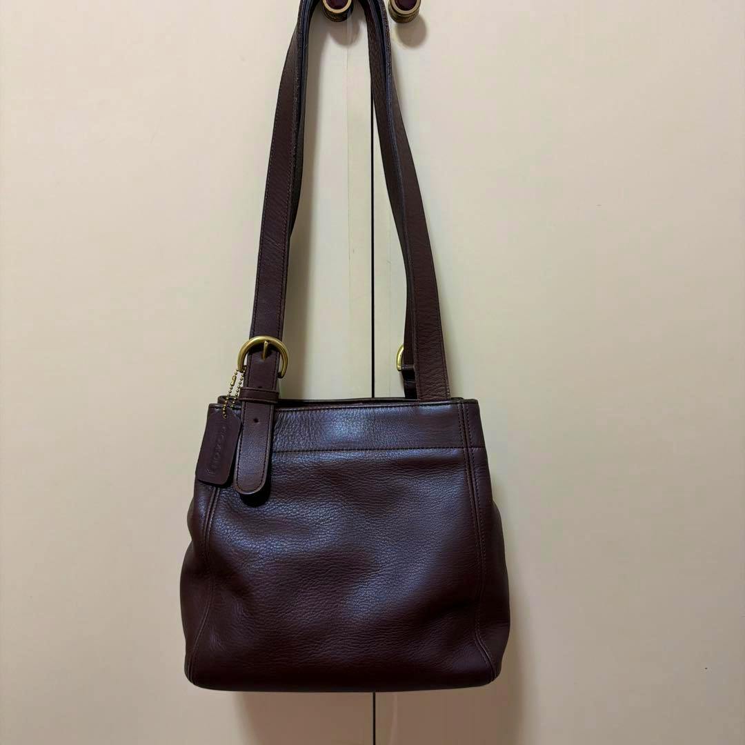 Front view of a brown OLD COACH cow leather shoulder bag hanging from two door knobs against a pale background.