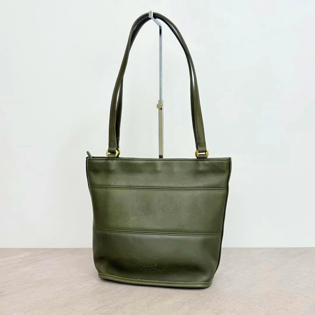 Front view of a khaki OLDCOACH cow leather shoulder tote bag hanging from a stand against a plain background.