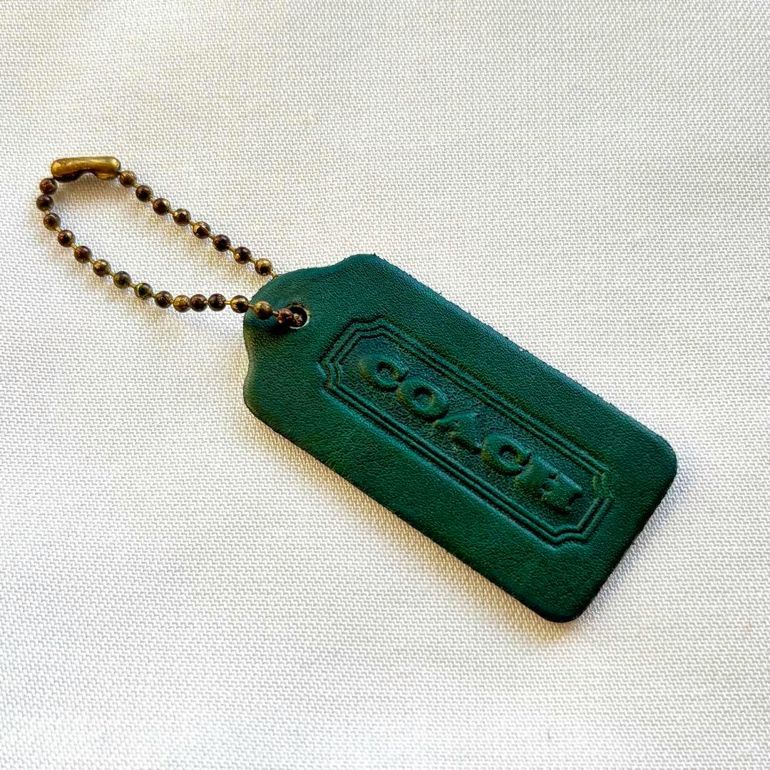 Green leather Coach keychain tag with a brass ball chain on a light textured background.