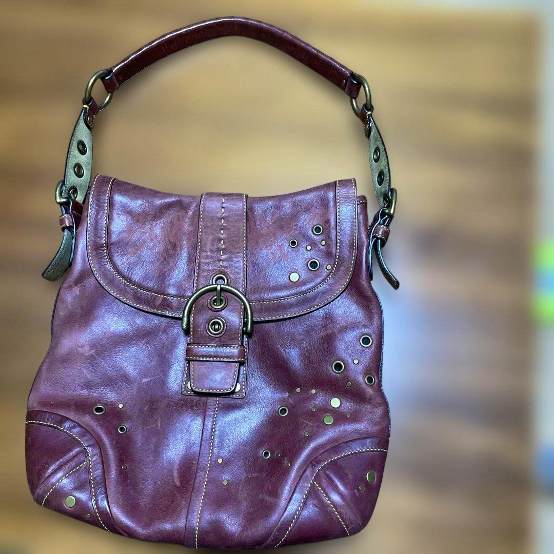 Burgundy OLD COACH leather shoulder bag with a buckle flap, grommet accents, and a short top strap against a blurred wooden background.