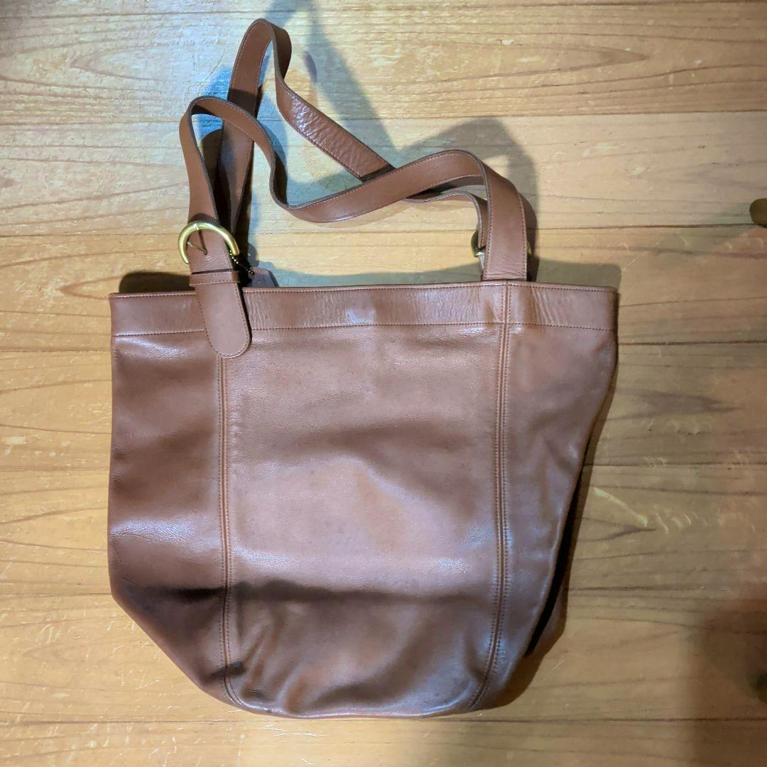 Brown leather shoulder bag laid flat on a wooden floor showing the front and straps.