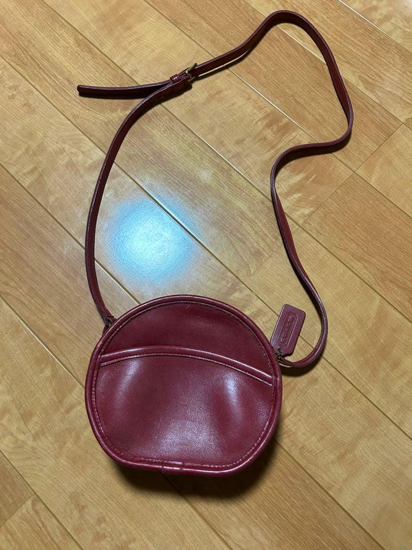 Red round leather shoulder bag with a long strap and hang tag placed on a wooden floor.