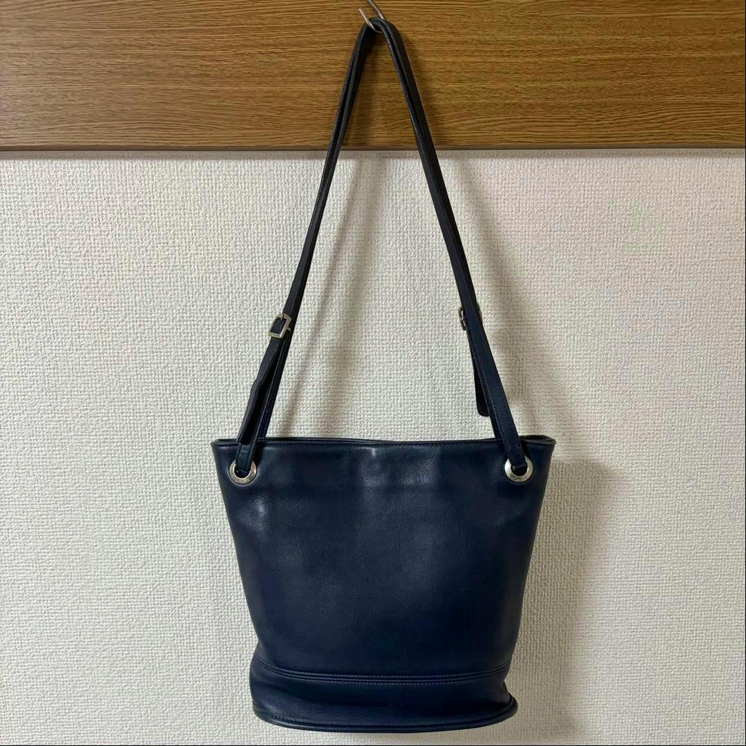 Navy bucket-style cow leather bag hanging on a hook against a textured wall.