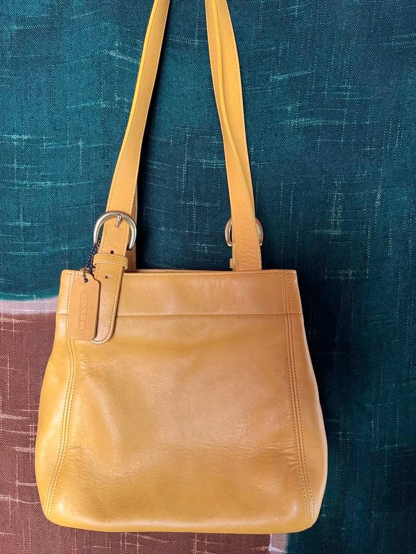 Yellow leather Coach shoulder bag hanging against a green and brown fabric background.