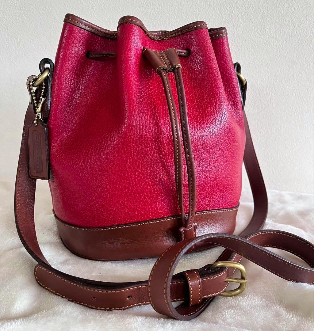 Red and brown Coach Sheridan drawstring bucket shoulder bag with brown leather strap and brass hardware on a light background.
