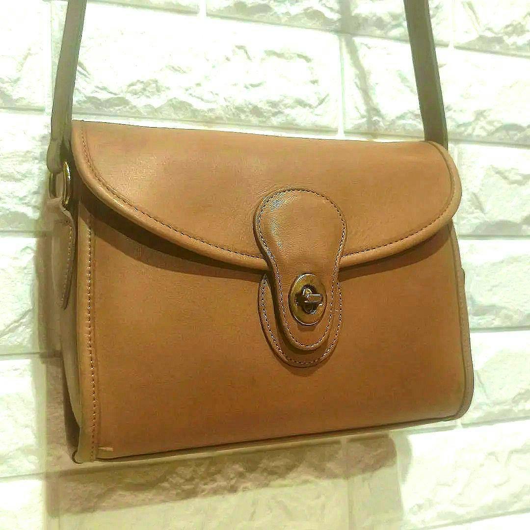 Camel leather Coach turn-lock shoulder bag hanging against a textured wall, front flap and turn-lock visible.