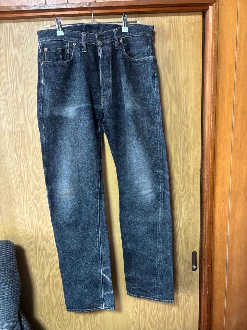Front view of the 旧DENIME black denim men's jeans hanging on a wooden door.