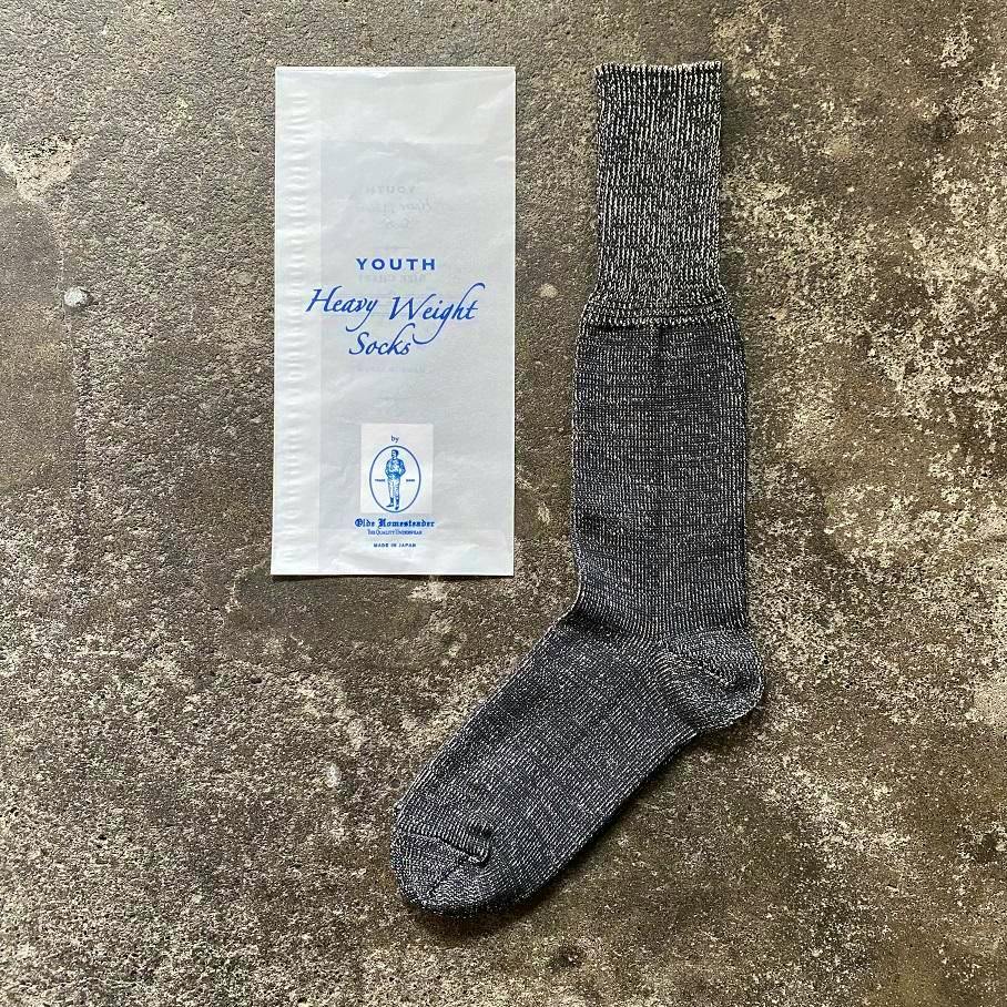 A single black marled crew sock displayed flat beside its white "Youth Heavy Weight Socks" packaging on a rough concrete surface.