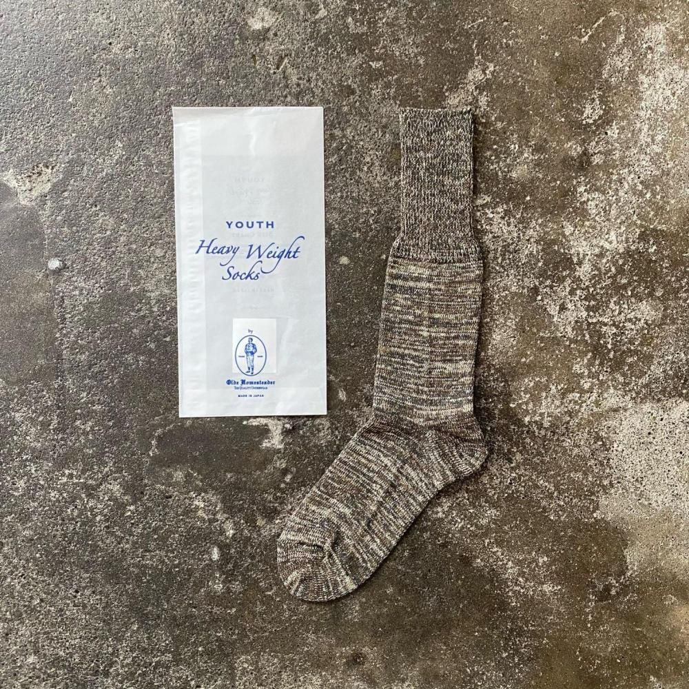 A single crew-length black-and-ecru tweedish jersey wool sock laid flat beside its white “Youth Heavy Weight Socks” bag on a textured surface.