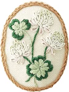 Oval embroidered brooch showing green clover motifs and white French-style stitched flowers on beige fabric with a braided border.