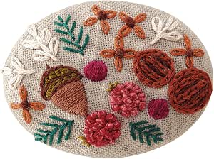 Oval embroidered brooch showing acorn and floral motifs in brown, pink, and green threads on beige fabric.