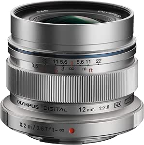 Three-quarter view of a silver OLYMPUS M.ZUIKO DIGITAL ED 12mm F2.0 camera lens showing focus and aperture markings.